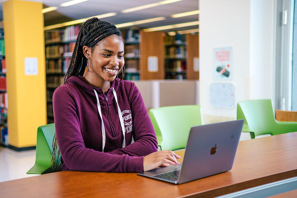 Student in a magenta hoodie smiling at a library table while working on her laptop.
