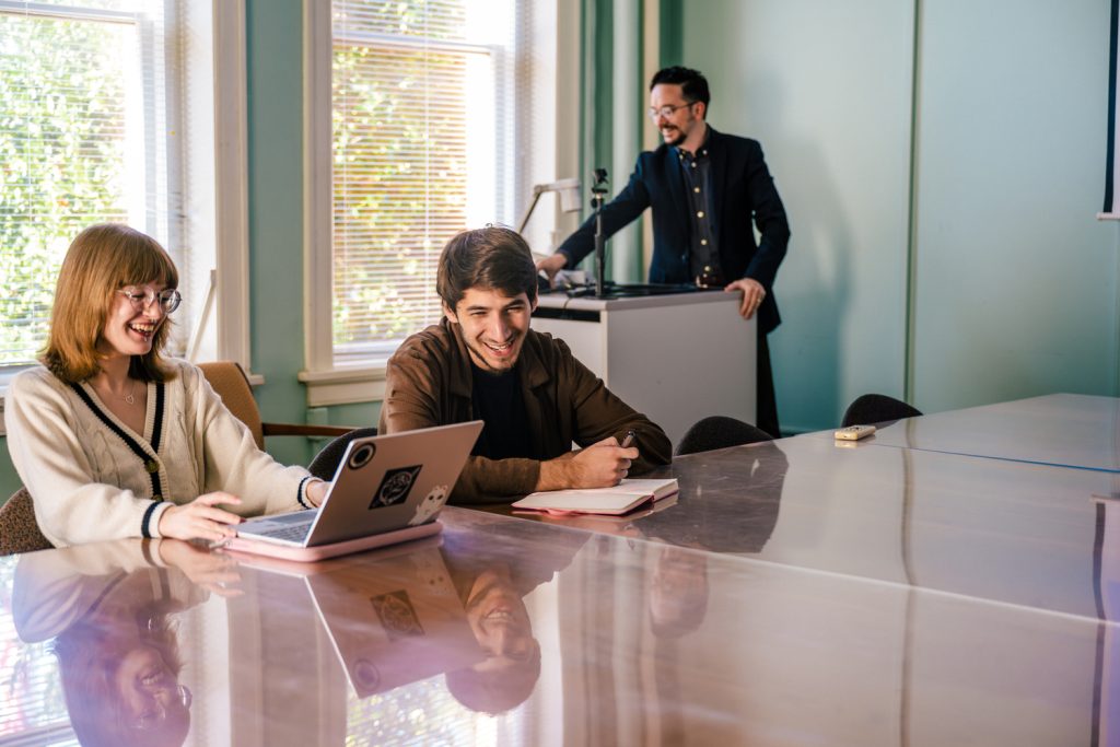 two students sitting at a conference table as the instructor stands at the front of the room